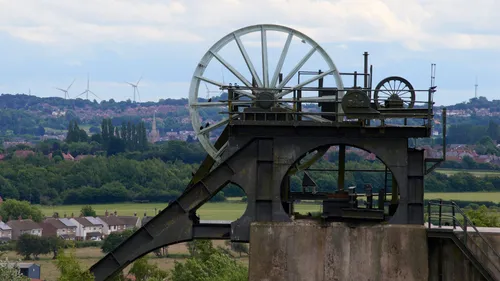 This landscape photograph, taken in late morning during the summer, prominently features the historic Pleasley Pit mining wheel in Derbyshire, United Kingdom. The mining heritage of the area is captured with the iconic wheel standing tall in the foreground, and modern wind turbines can be seen scattered across the countryside in the background, demonstrating a contrast between traditional industry and renewable energy. Solar panels are also visible, further highlighting this blend of old and new energy sources. The village of Pleasley, with its distinctive church spire rising amongst the trees and houses, adds a picturesque touch to the rural Derbyshire scenery. The photograph effectively encapsulates the region’s mining history while also showcasing the shift towards sustainable energy in the United Kingdom.